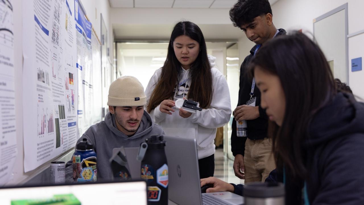 Four students work together in a hallway with laptops and scientific posters on the wall; one student holds a device while others type or observe.