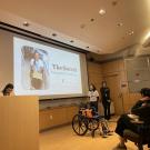 A group presents "The Swivel Completed Prototype" in a lecture hall; a slide shows a person using the device and a wheelchair with an attached tray is displayed in front.